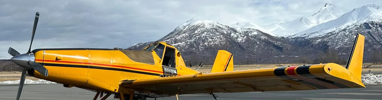 A yellow crop duster aircraft with black and red stripes on an airfield, with snowy mountains in the background.