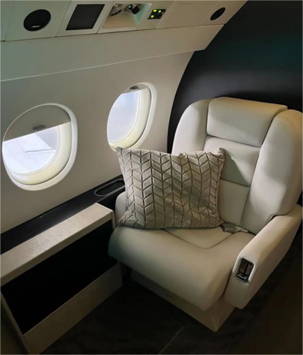 A close-up of a plush white leather club seat with a patterned throw pillow next to two oval windows in the aircraft cabin.