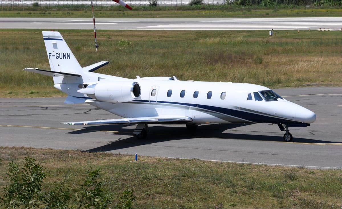 A white Cessna CitationJet with blue and grey stripes taxiing on an airport runway.