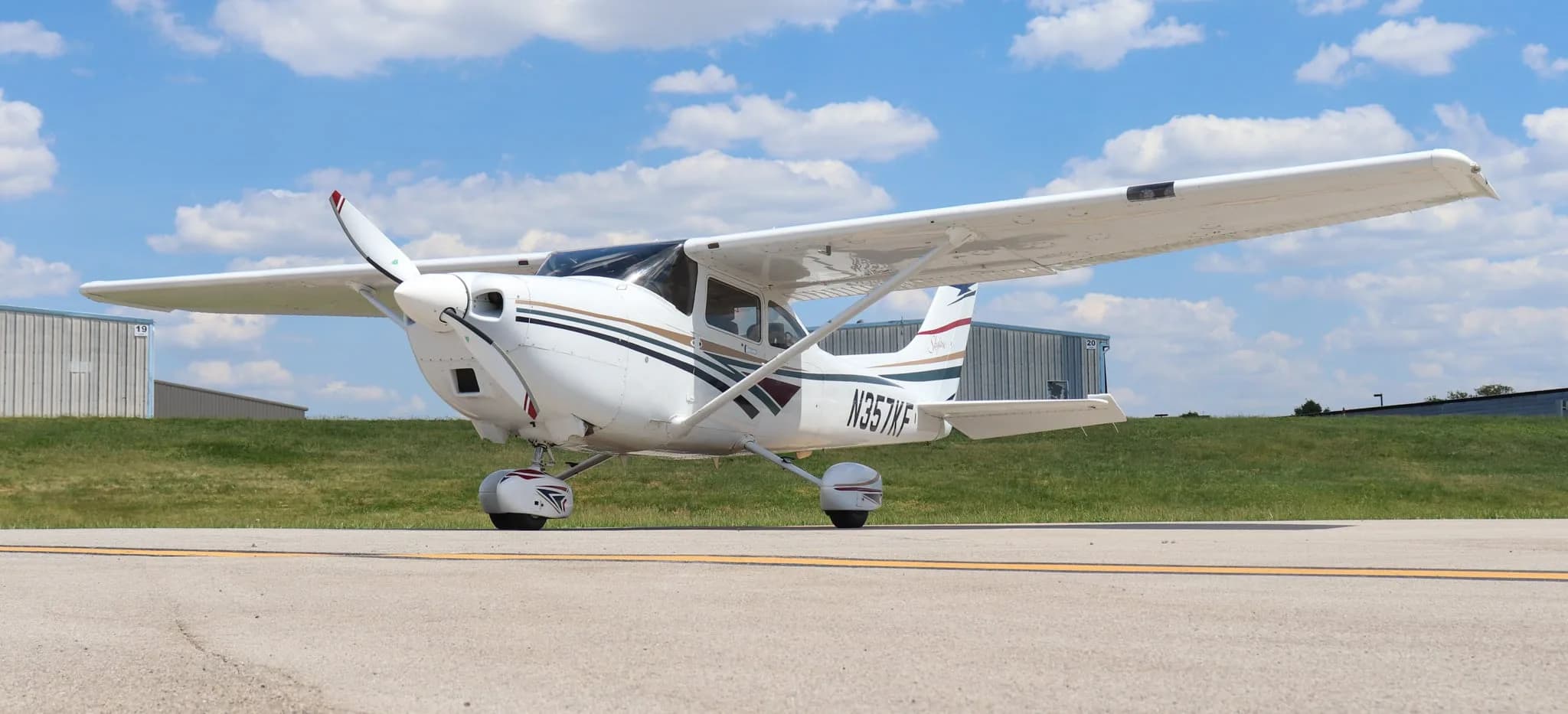 A white Cessna 182 Skylane with gold, green, and maroon stripes parked on an airport ramp under a blue sky.