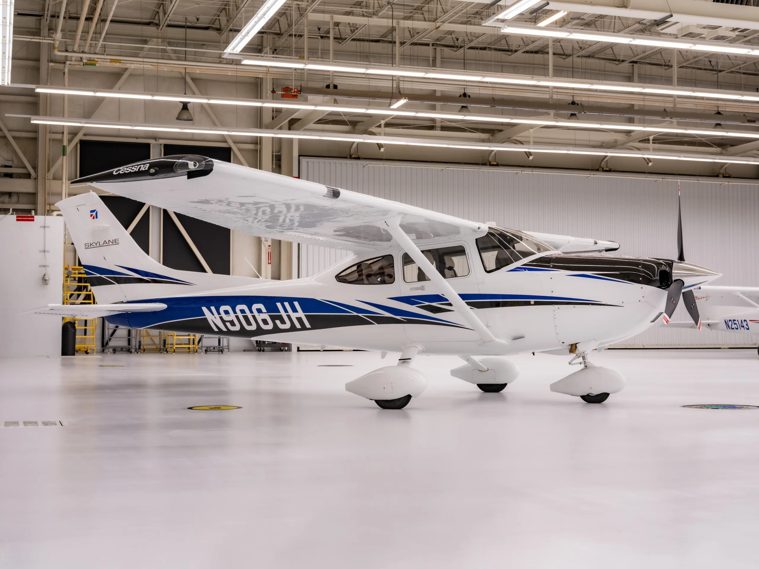 A white Cessna Skylane N906JH with blue and black accents is parked in a brightly lit hangar.
