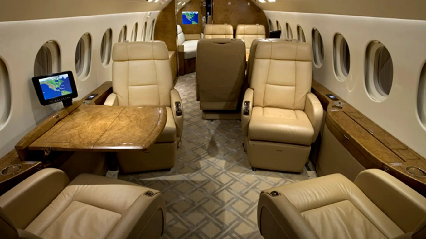A luxurious aircraft cabin interior with beige leather seats, a wooden table, and small display screens.