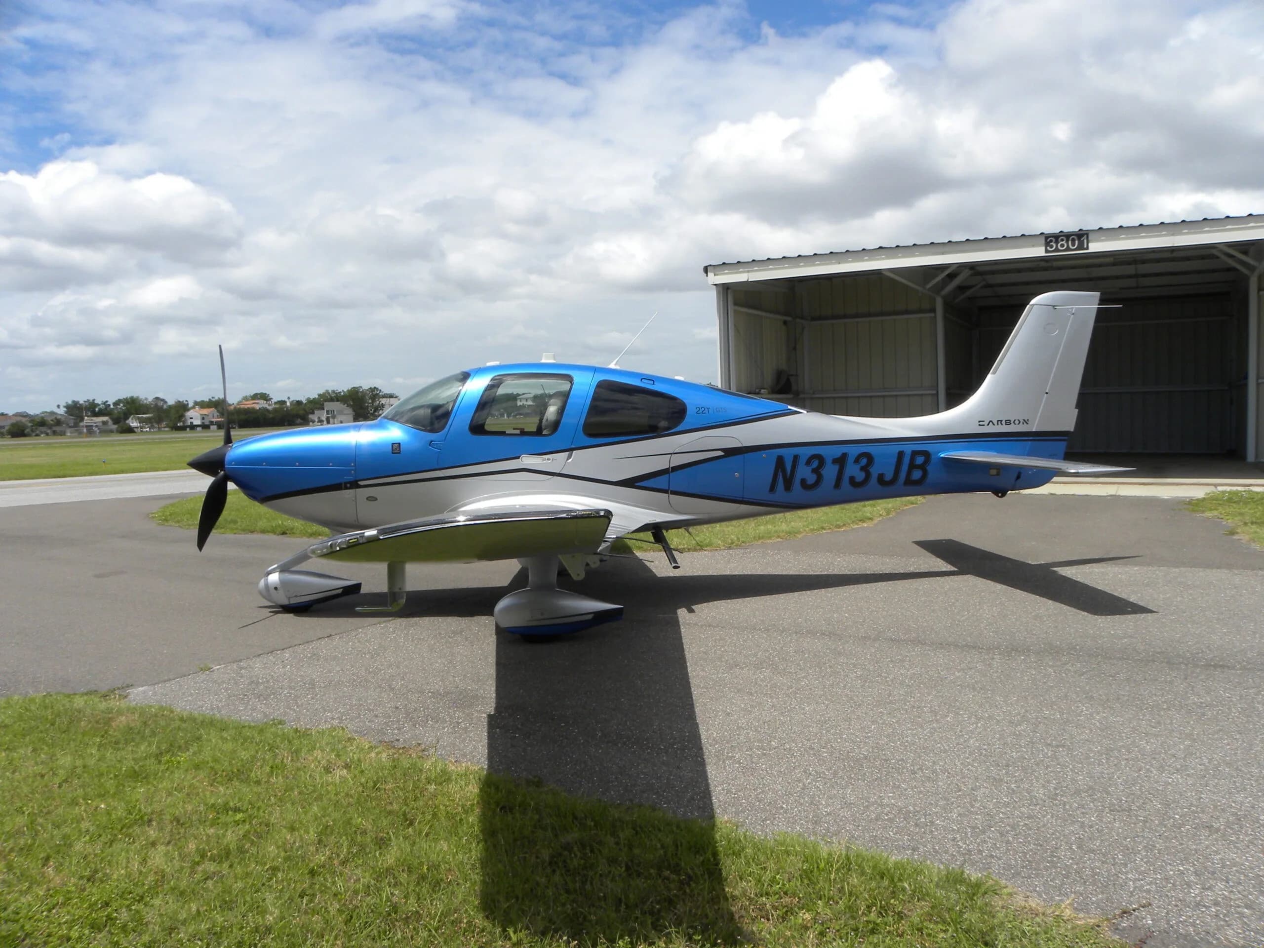 A blue and silver Cirrus SR22T aircraft, N313JB, parked on an airport tarmac next to a hangar.