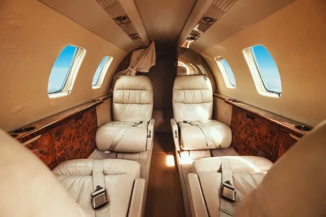 The interior cabin of a business jet featuring light beige leather seats and wood-grain paneling.