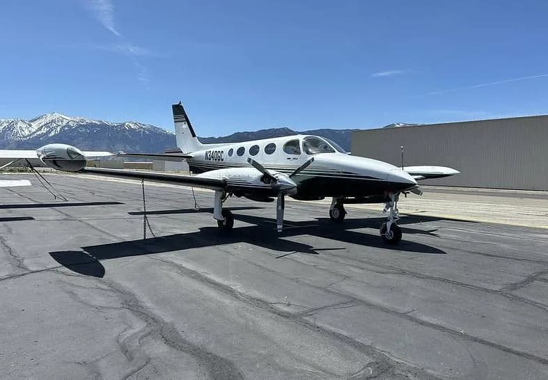 A white and dark green twin-engine Cessna 340A (N340GC) is parked on an asphalt ramp with snow-capped mountains in the background under a clear blue sky.