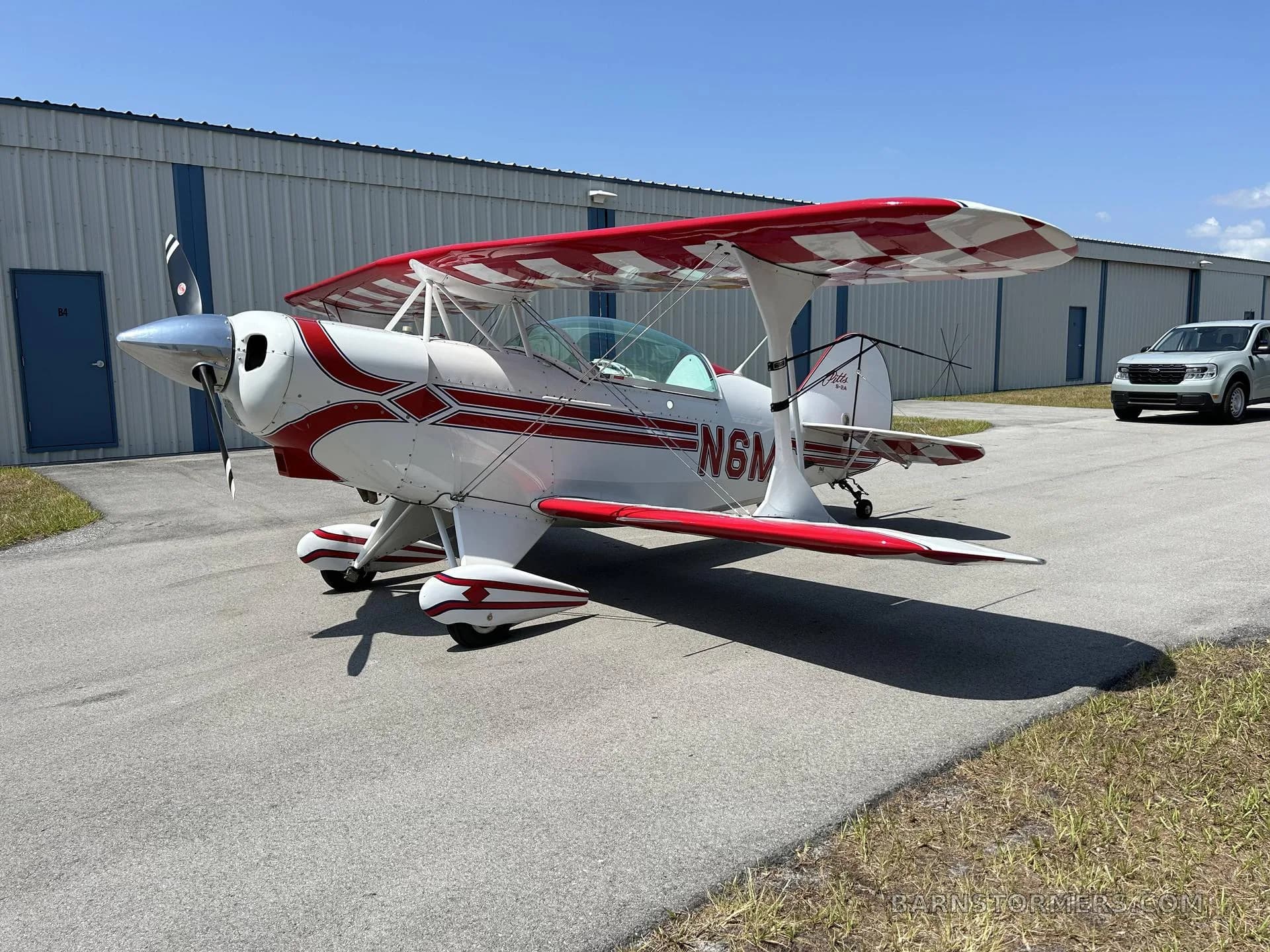A white Pitts Special biplane with red checkered wings and red stripe accents, parked on an asphalt ramp with a white truck in the background.