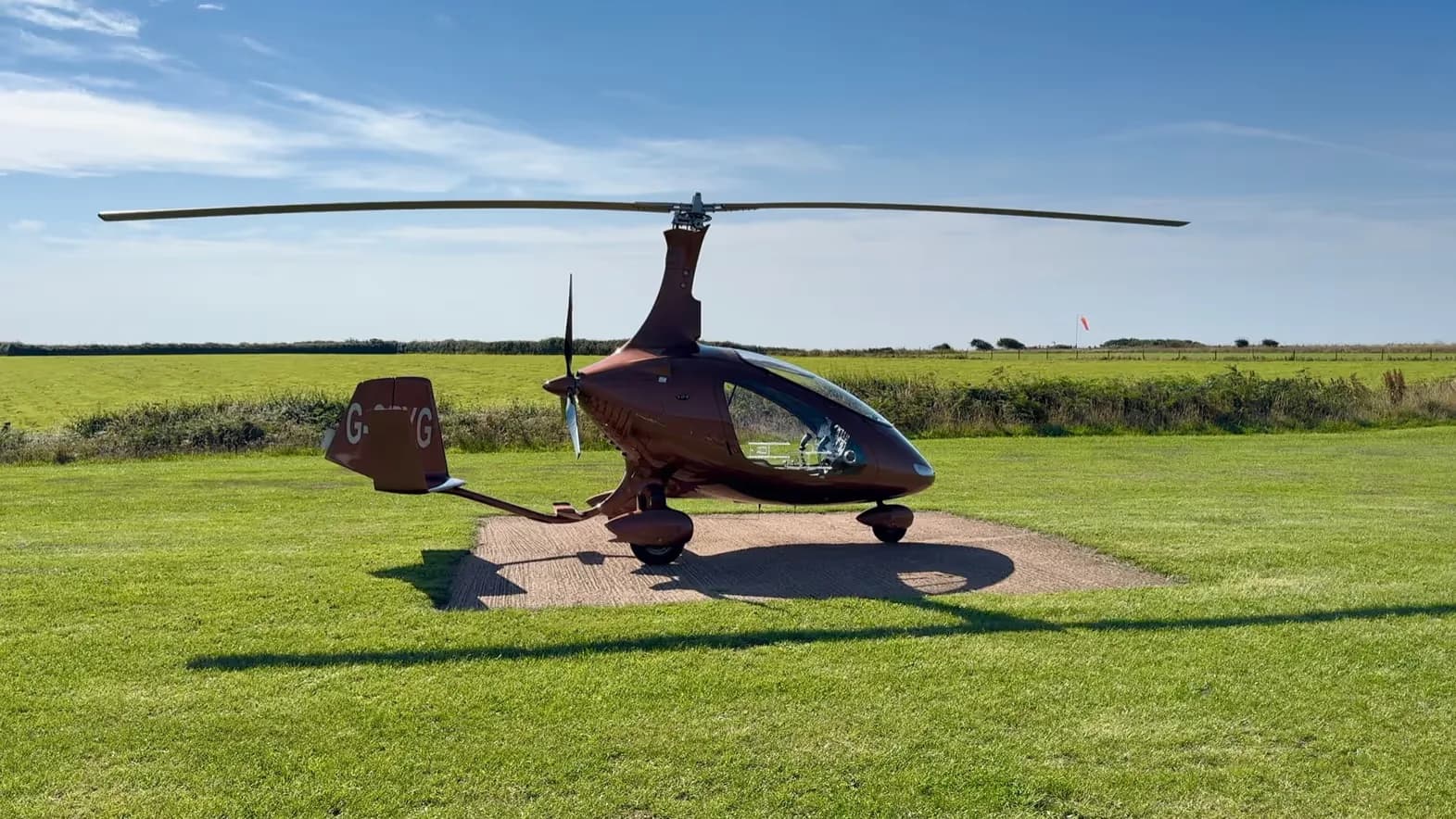 A brown gyrocopter, G-ORYG, parked on a concrete pad in a grassy field under a blue sky, viewed from the front-right.