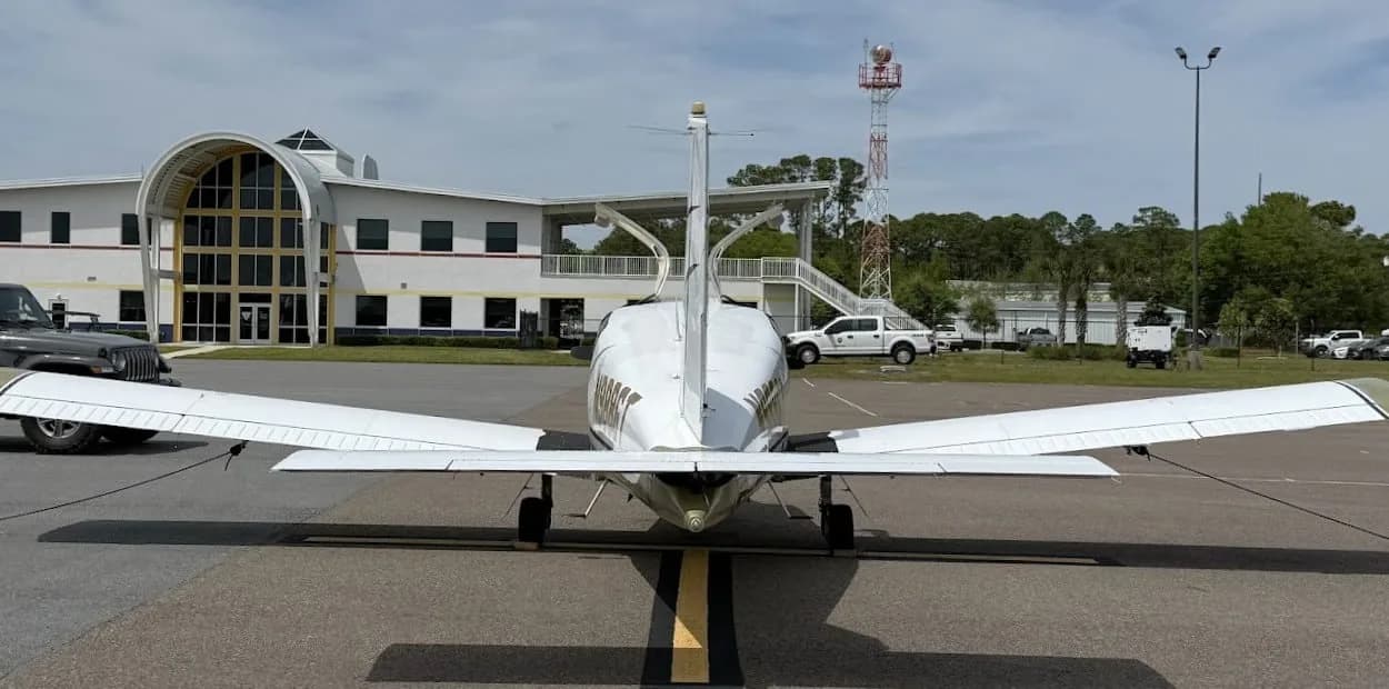A rear view of the white and gold Socata TB-20 Trinidad aircraft with its doors open, parked in front of an airport building.