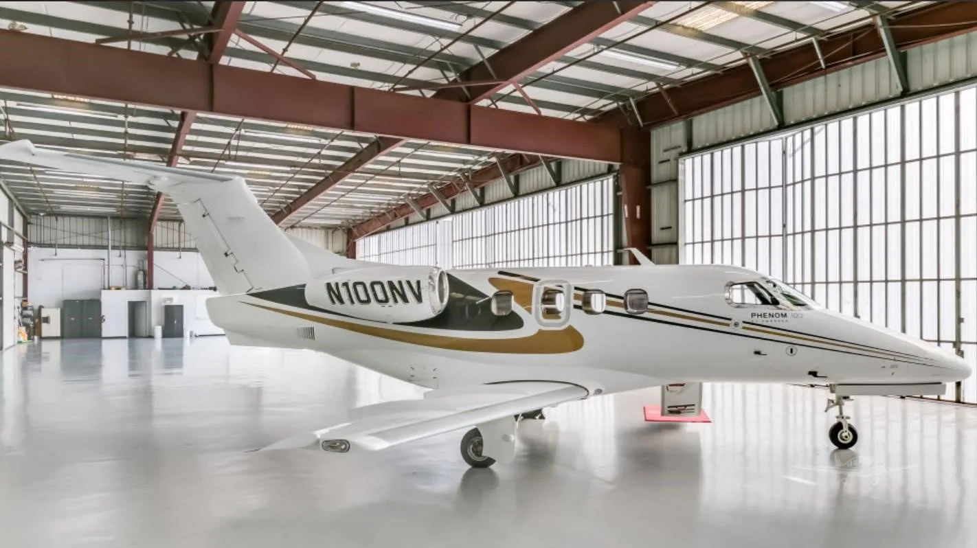 A white Embraer Phenom 100 with gold and black stripes (tail number N100NV) parked inside a hangar.