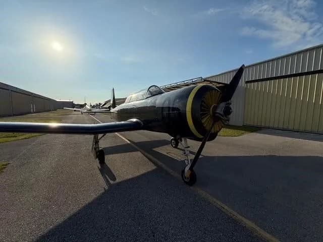 A low-angle front-right view of a dark gray Yak-52 aircraft with a yellow propeller spinner, parked on an asphalt ramp.