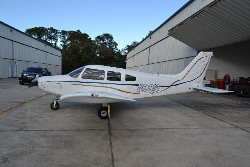 Side view of a white and silver Piper PA-28-181 Archer II aircraft, N6789T, parked in front of a hangar.