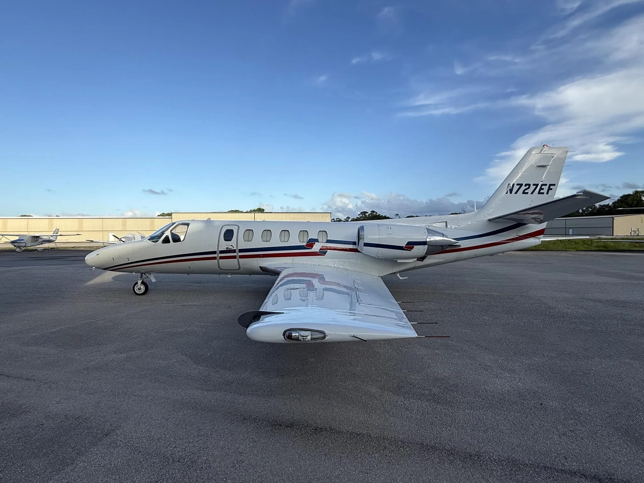 A white Cessna Citation business jet (N727EF) with red and blue stripes parked on an airfield, showing the right side.