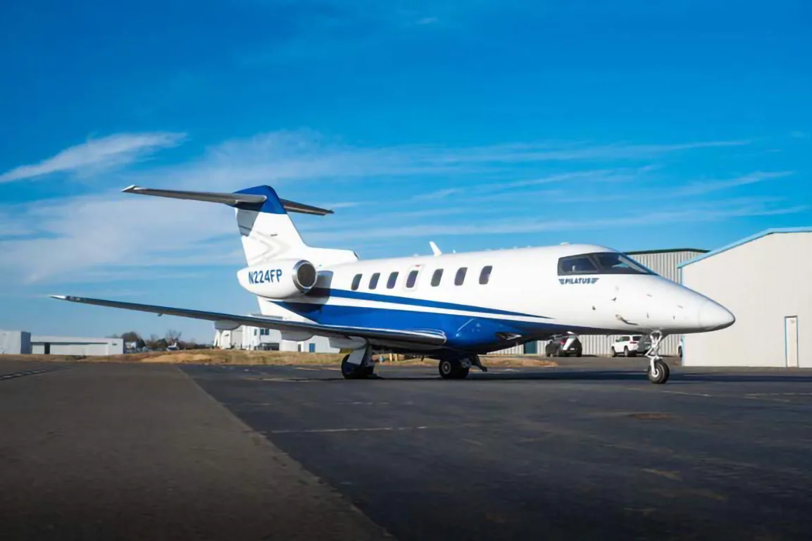 A Pilatus PC-24 jet with tail number N224FP, painted white and blue, is parked on an airfield next to a hangar.