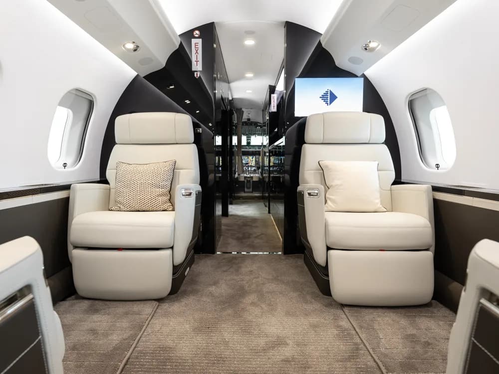 The interior of a private jet cabin with two light grey seats in the foreground and a screen displaying a logo at the rear.