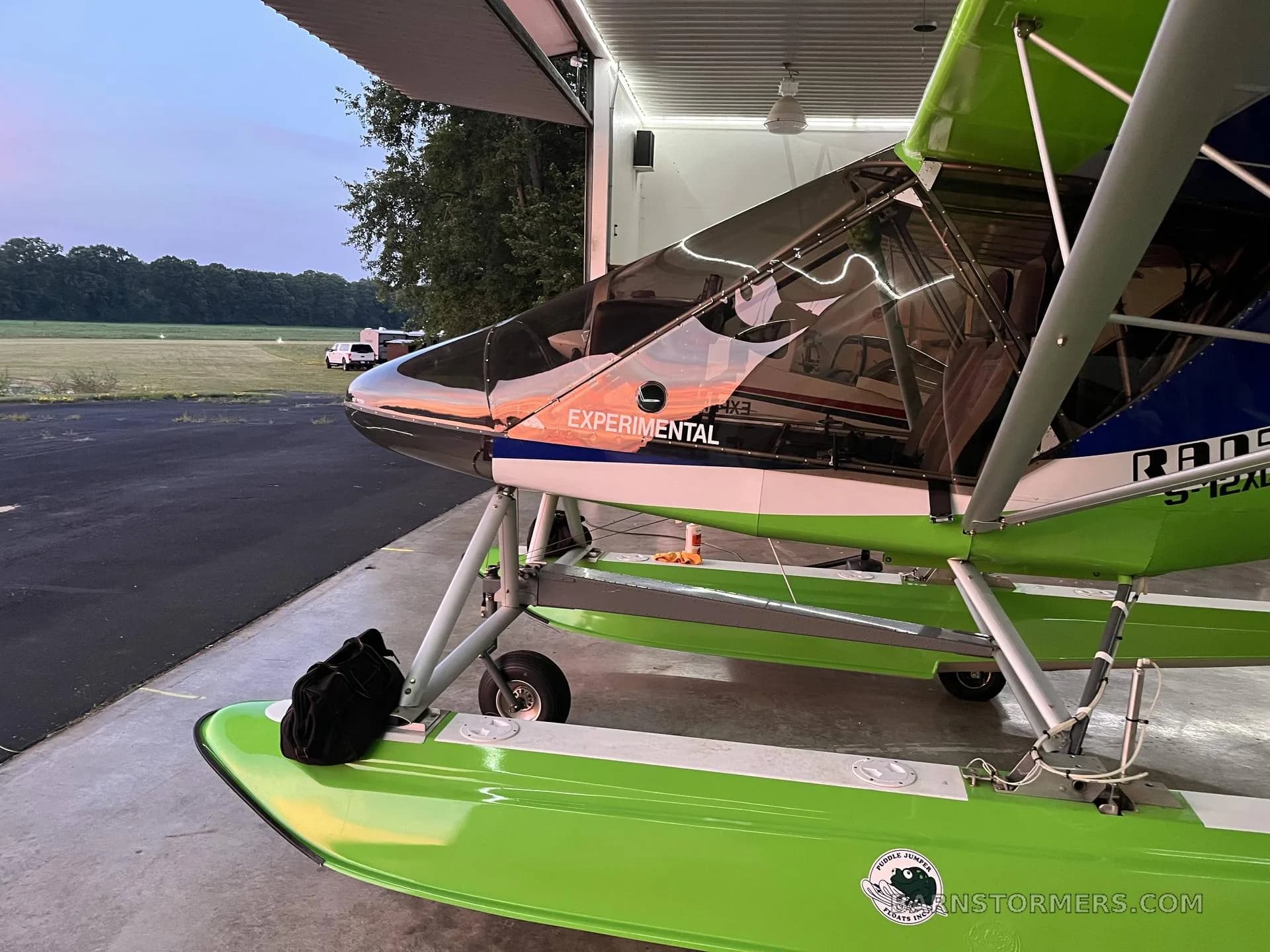 The front of a bright green and blue ultralight seaplane with floats is parked inside a hangar at dusk, reflecting the sunset.