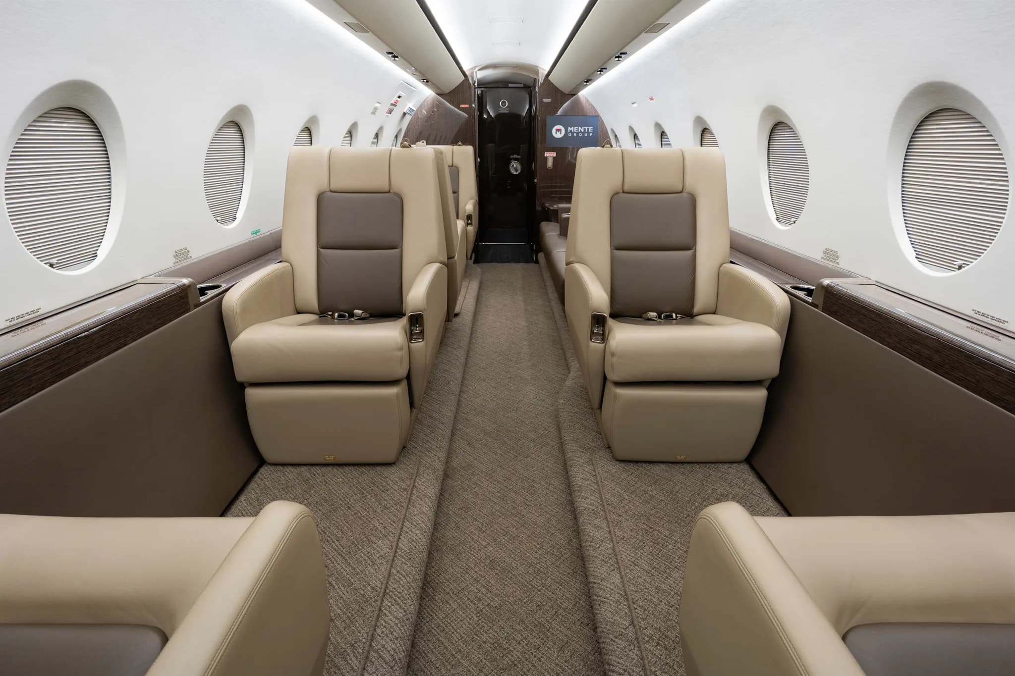 The interior of a private jet cabin, showing an aisle between two rows of comfortable beige armchairs.