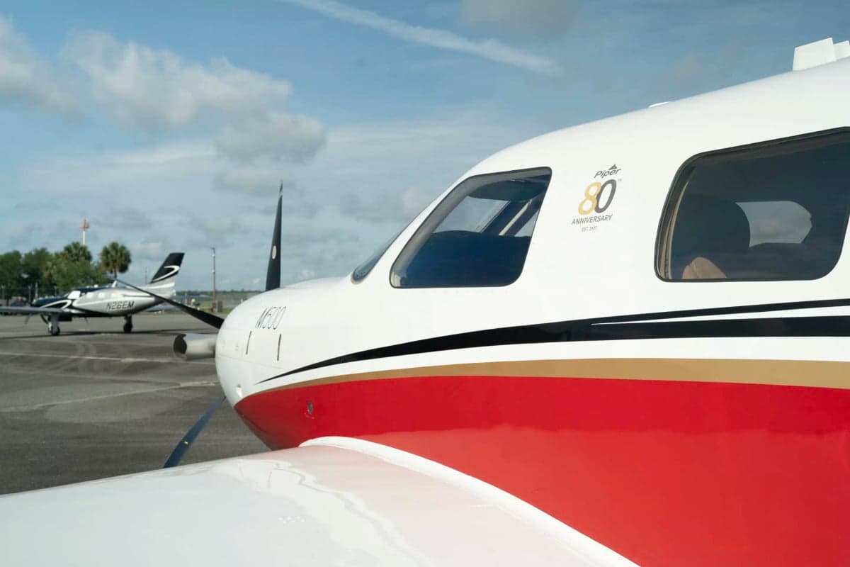 A close-up of the cockpit area of a white and red Piper M500, showing "Piper 80th Anniversary" text on the fuselage.