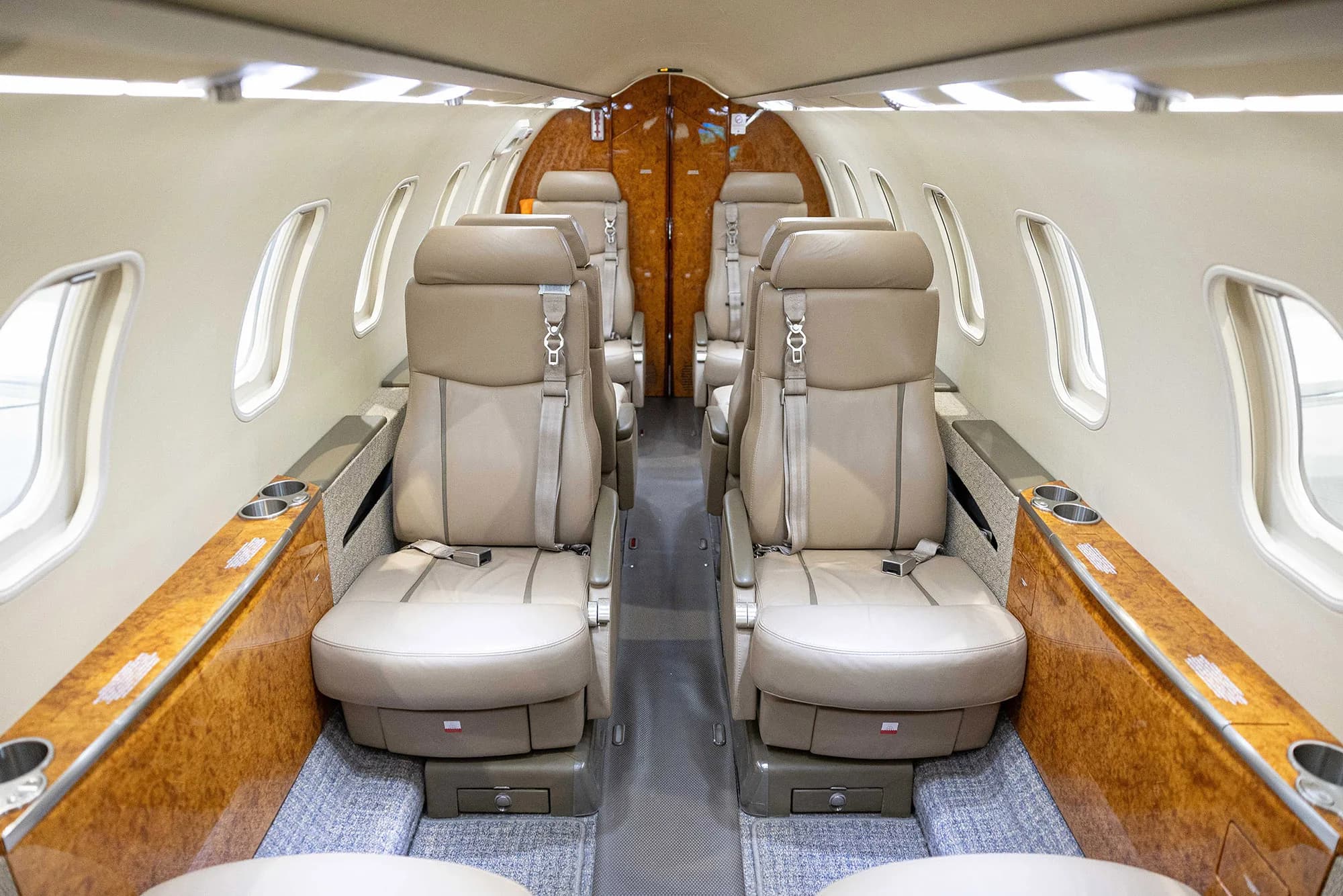 The luxurious interior cabin of a private jet with tan leather seats and polished wood paneling, viewed from the front.