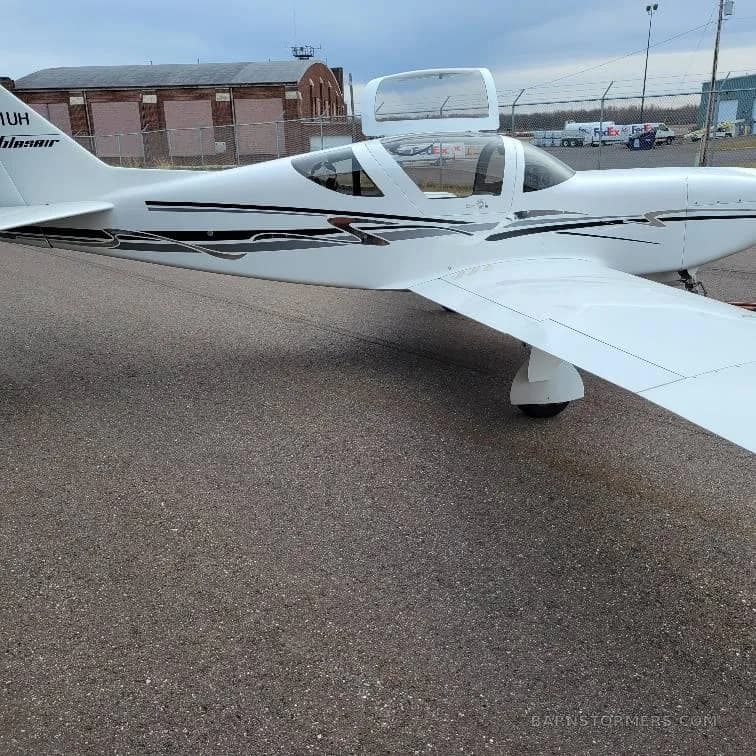 A white Vans RV-10 or similar kit plane with black and grey stripes, parked on an asphalt ramp with its canopy open, showing the left side.