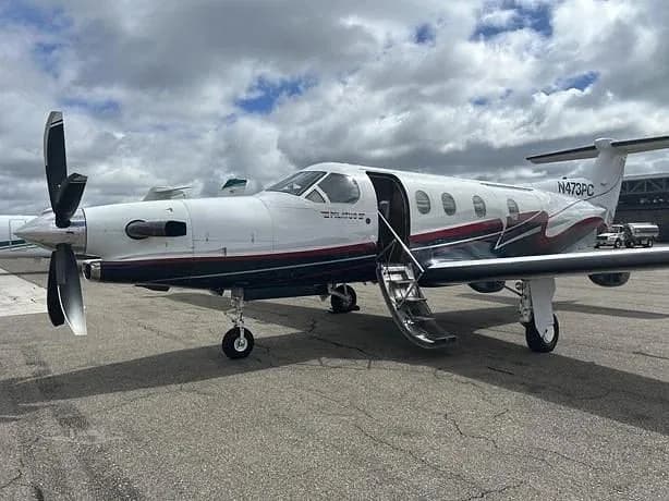 A white Pilatus PC-12 with dark blue and red stripes is parked on an airport tarmac under a cloudy sky, with its passenger door open.