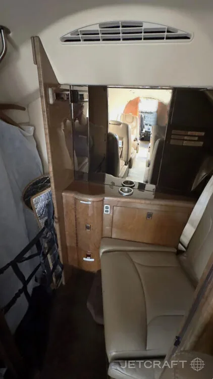 A view inside the business jet cabin, showing a tan leather seat, a wooden cabinet with a mirror, and a glimpse of the aft cabin.
