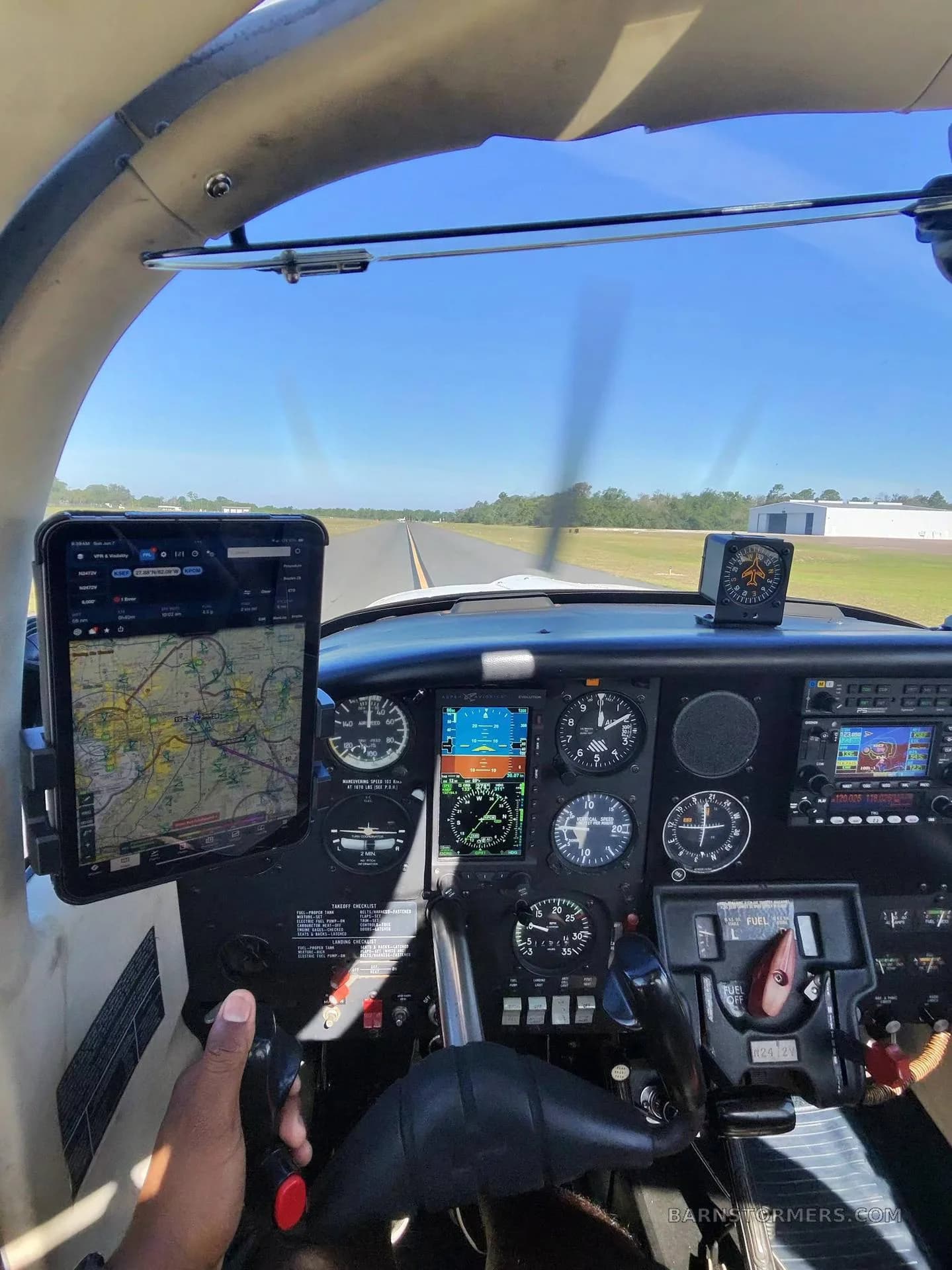 View from a cockpit showing the instrument panel, a pilot's hand on the yoke, and a runway visible through the windshield.