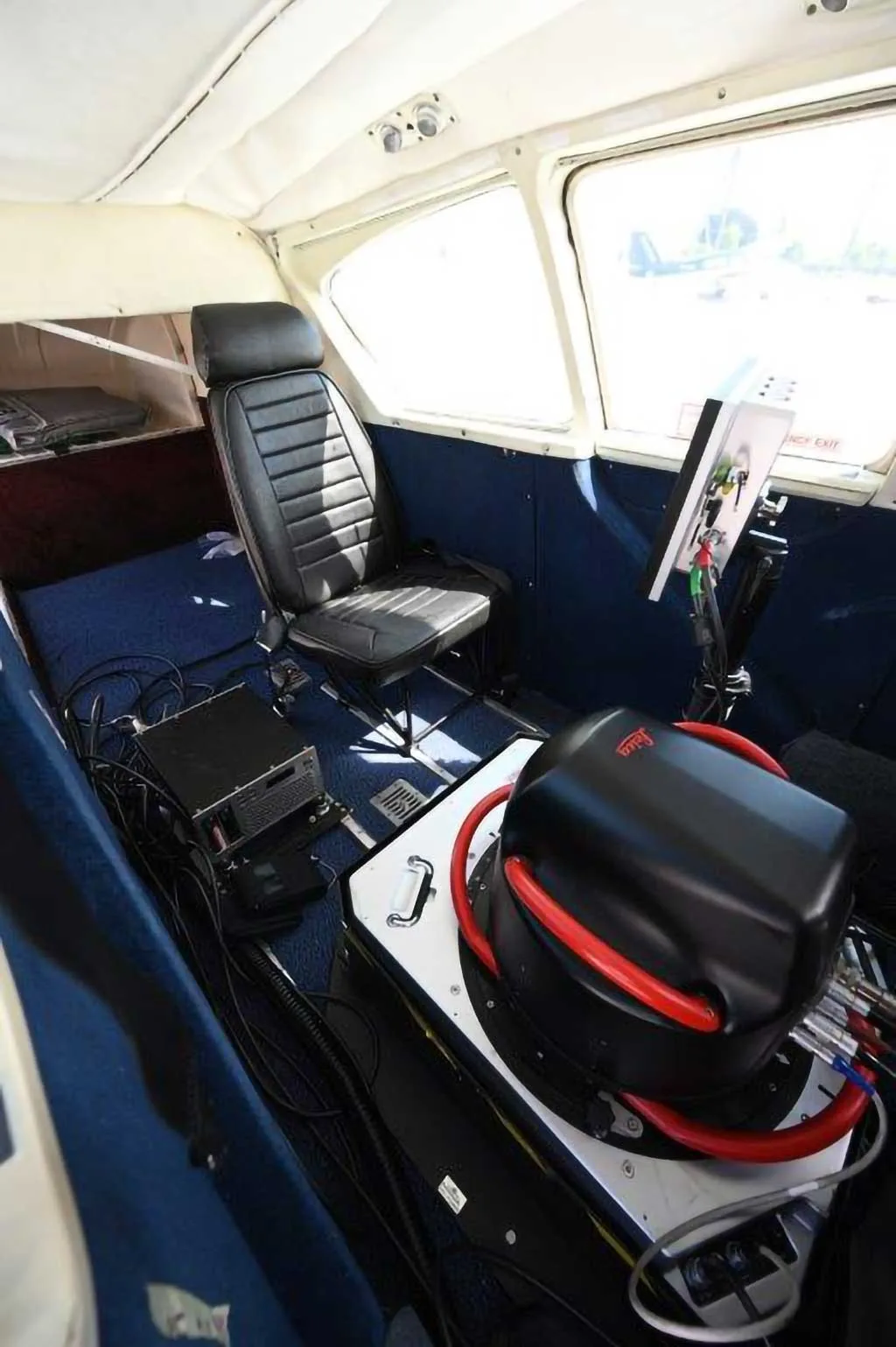 The interior of an aircraft cabin showing a black seat, blue carpet, and a black Leica sensor unit with red tubing, looking towards the front.