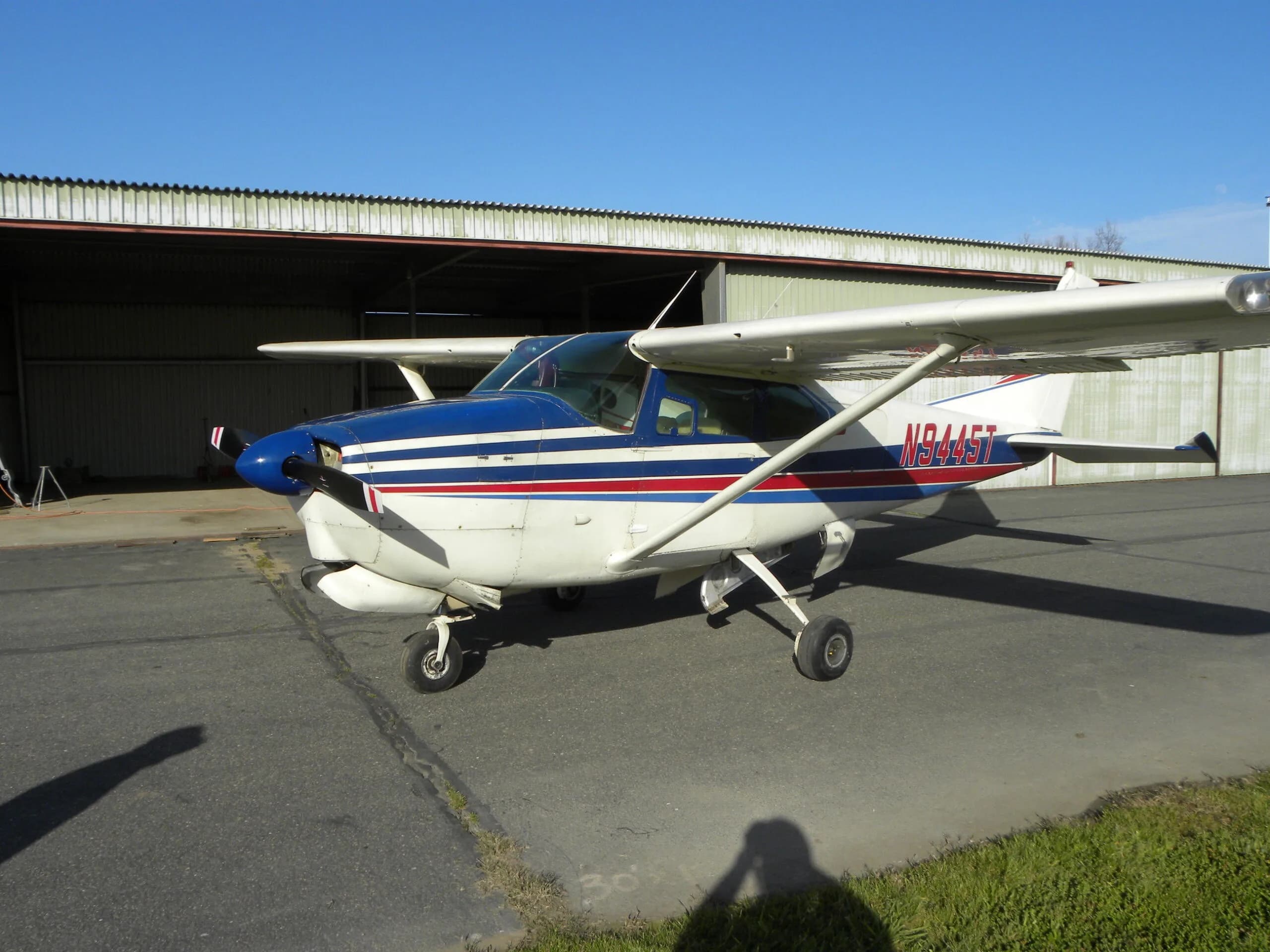 A front-side view of a white Cessna 182 with blue and red stripes, registration N9445T, parked near a hangar.
