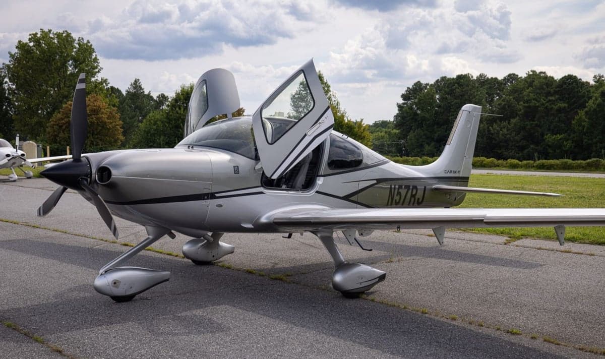 A silver Cirrus SR22 aircraft (N57RJ) with black accents, parked on an airfield with its gull-wing doors open.