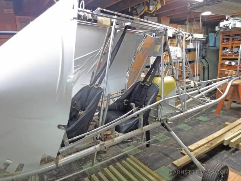 A side view of a partially assembled ultralight aircraft frame with two black seats and white fabric in a workshop.