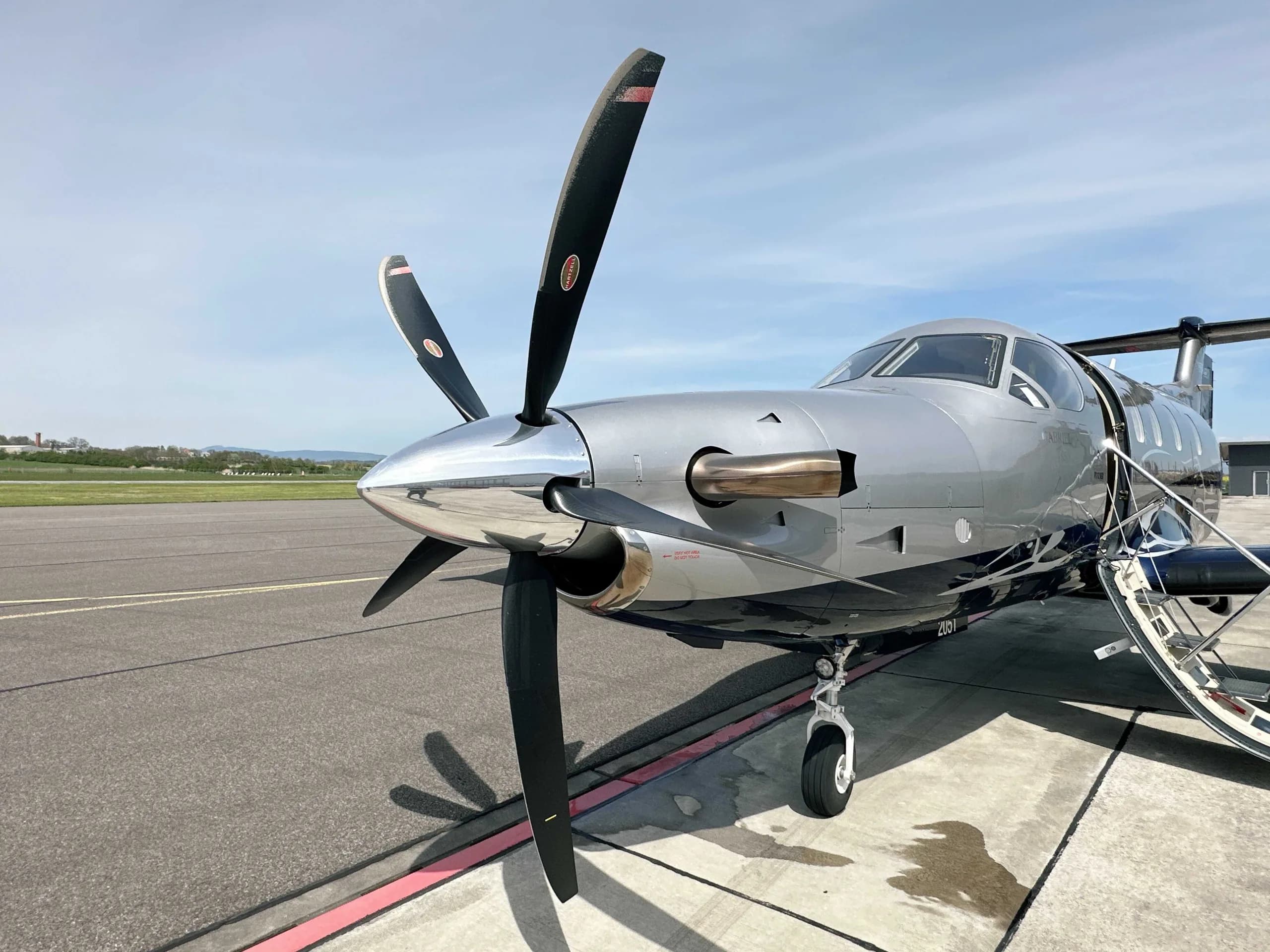 A close-up view of the nose and propeller of a silver and blue Pilatus PC-12 NGX aircraft on an airport tarmac.