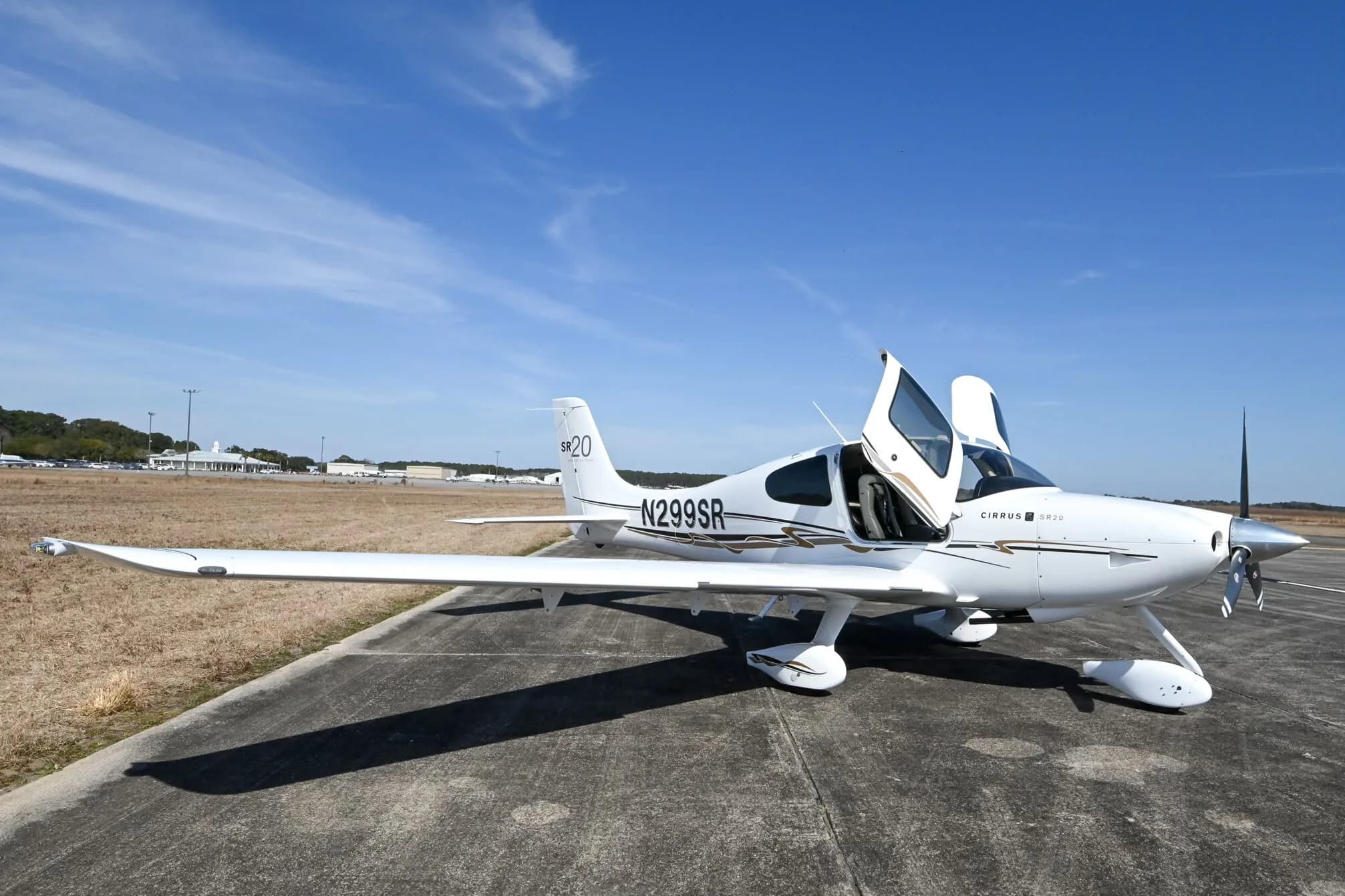 A white Cirrus SR20 aircraft with black and gold accent stripes, registration N299SR, parked on an airfield with both doors open.