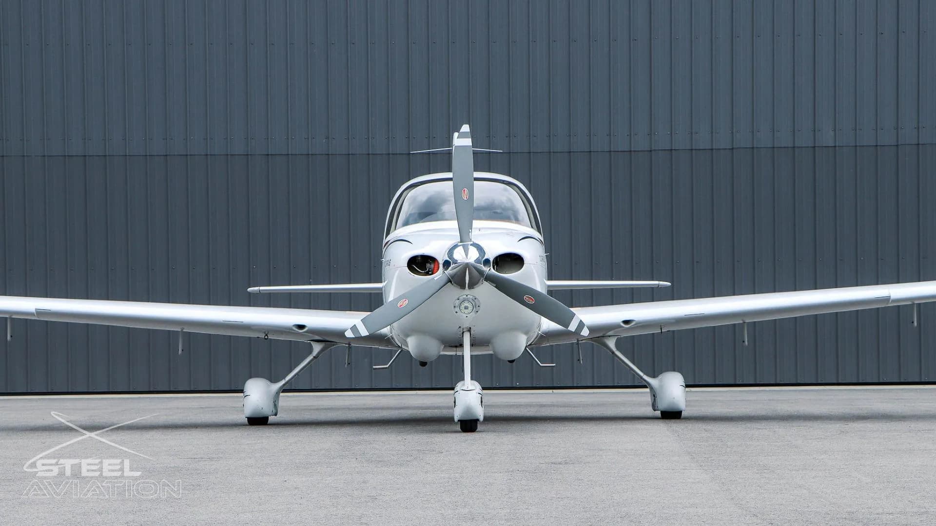 A close-up front view of a white Cirrus SR22 aircraft with a dark grey hangar in the background.