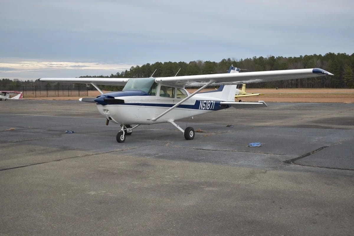 A white Cessna 172 with blue and grey stripes is parked on a tarmac, viewed from the front-right.