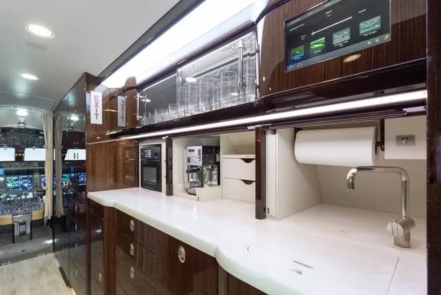 The well-equipped galley of a business jet, featuring a sink, microwave, coffee maker, and an entertainment screen.