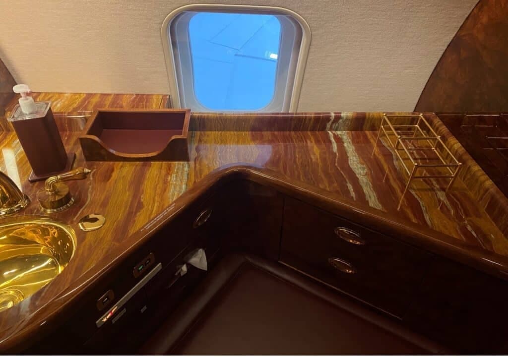 A close-up of a lavatory or galley counter with a golden sink, marbled brown countertop, and a window.