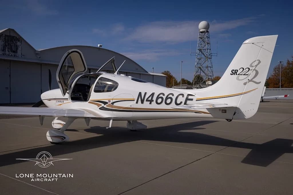 A white Cirrus SR22 aircraft with gold and black stripes, registration N466CE, parked on an airfield with its doors open, viewed from the rear-right.