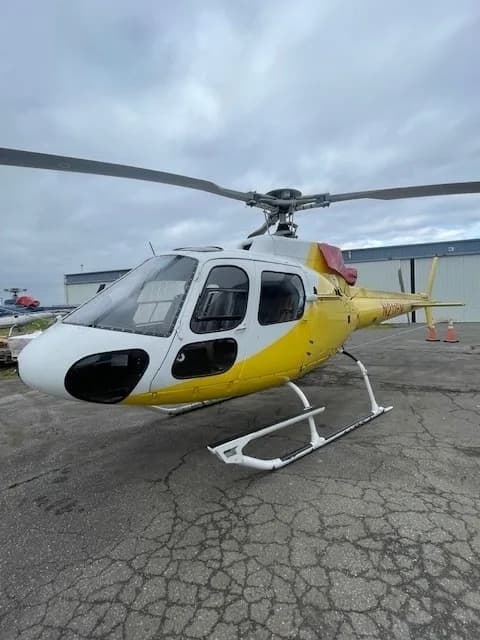 A white and yellow Eurocopter AS350 B3 helicopter, N211FN, is parked on an asphalt tarmac under a cloudy sky.