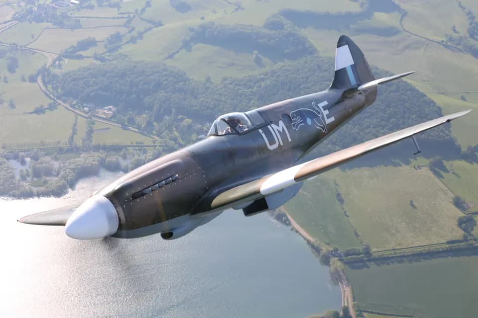 A Supermarine Spitfire in flight, banking over a lake and rolling green hills.