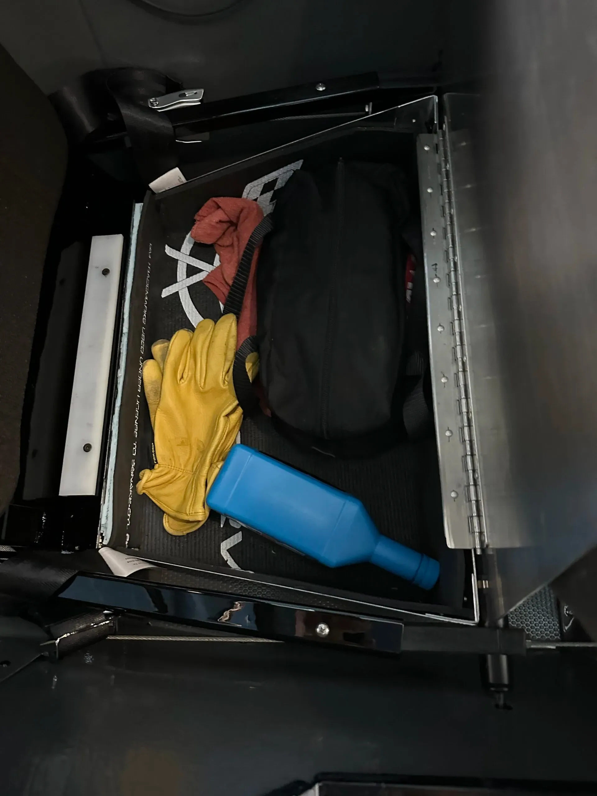 An open storage compartment in the aircraft, containing yellow gloves, a black bag, and a blue bottle.
