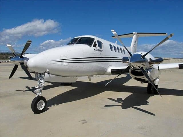 A white Beechcraft King Air with black stripes parked on an airport tarmac under a blue sky with scattered clouds.