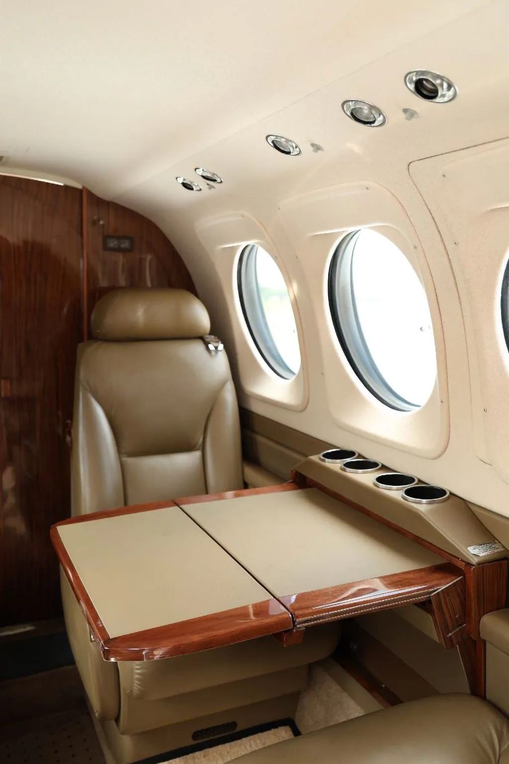 An aircraft interior showing a tan leather seat and a fold-out table with cup holders next to a window.