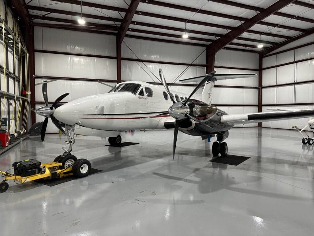 A white Beechcraft King Air with black and red stripes parked in a hangar, viewed from the front left.