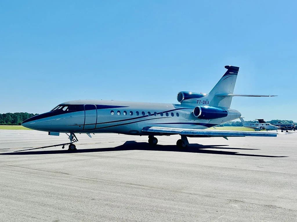 A Dassault Falcon 900 business jet with blue and white livery is parked on an airfield under a clear sky.