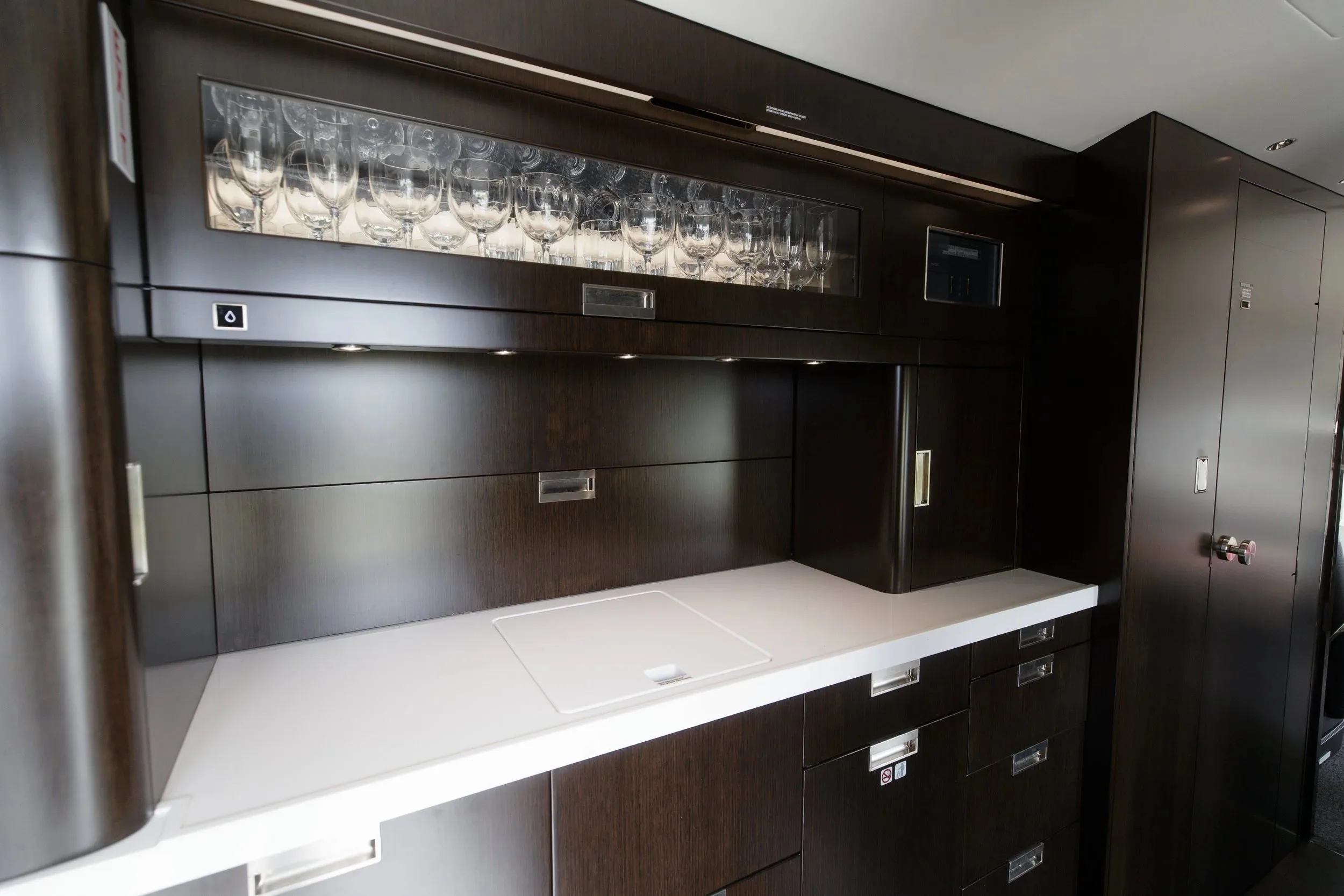 A luxurious aircraft galley features dark wood cabinetry, a white countertop, and a display of glassware.