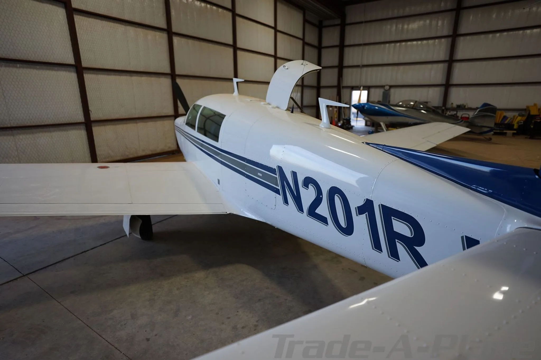 An overhead rear view of the white aircraft N201RJ with blue and grey stripes, parked in a hangar with its door open.