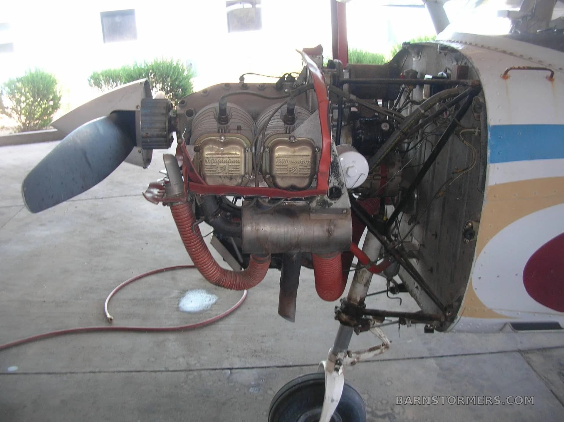 The exposed engine of a white aircraft, showing the cylinders and exhaust system, with the propeller visible on the left.