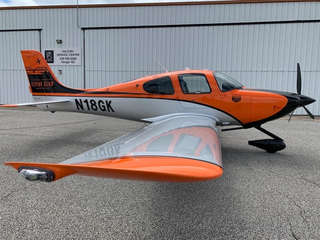 Side view of a Cirrus SR20 aircraft with orange, black, and silver paint scheme, parked on an airfield.
