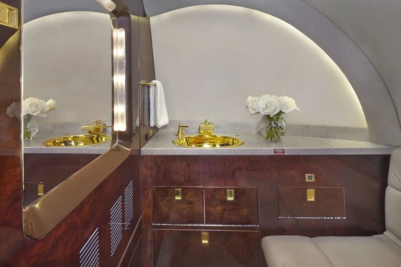 A luxurious aircraft lavatory with a gold sink, dark wood cabinetry, and a large mirror with vertical lighting.