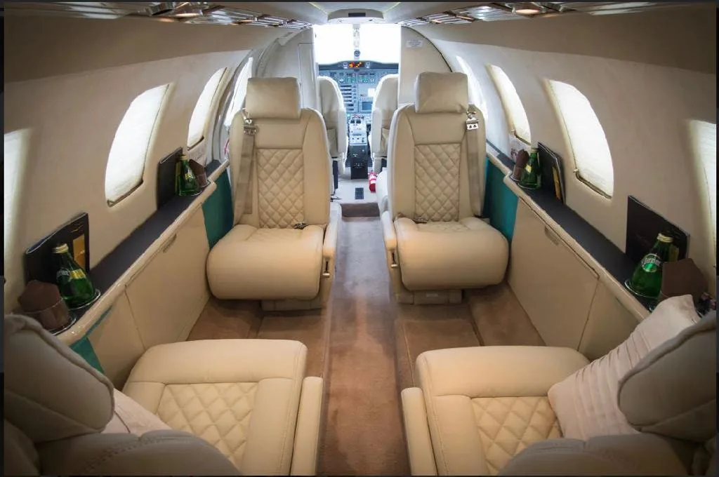 The luxurious cabin interior of a private jet with quilted beige leather seats and a view into the cockpit.